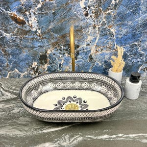 Puede incluir: Lavabo ovalado de cerámica con un patrón geométrico en blanco y negro. El interior del lavabo es de color crema con un diseño floral alrededor del desagüe. Un grifo dorado está montado encima. Un dispensador de jabón y un jarrón pequeño.