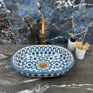 Puede incluir: Lavabo ovalado de cerámica con un patrón geométrico azul y blanco. El lavabo tiene un desagüe de latón y está sobre una encimera oscura y una pared con aspecto de mármol azul. Se ve un grifo dorado.