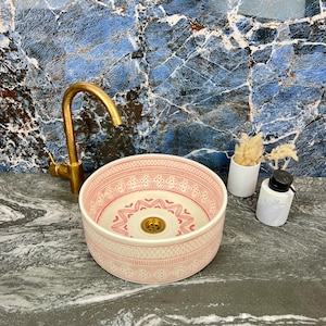 Puede incluir: Un lavabo redondo de cerámica rosa y blanco con un intrincado diseño. Un grifo dorado está montado a la izquierda. Un dispensador de jabón blanco y un pequeño jarrón con flores secas están a la derecha. El lavabo está sobre una encimera gris.