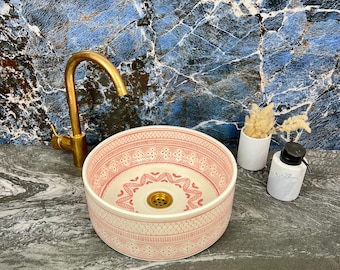 May include: A round, pink and white ceramic sink with a detailed pattern. A gold tap is mounted to the left. A white soap dispenser and small vase with dried flowers are to the right. The sink is on a grey countertop.