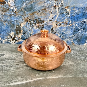 May include: A copper-colored, hammered metal serving dish with a matching lid. The dish has two handles and a decorative knob on top of the lid. The surface has a dimpled texture. The background is a blue and white marbled pattern.