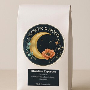 Puede incluir: Una bolsa blanca de café en grano entero con un logotipo negro y dorado que presenta una luna creciente y una flor roja. La bolsa está etiquetada con "FLOWER & MOON Obsidian Espresso Tostado oscuro Chocolate negro, azúcar moreno, canela Café en grano entero."