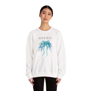 Pode incluir: Camisola branca com laço azul e design floral. O texto "YOUR TEXT" está impresso acima do laço. Uma peça de vestuário de estilo casual.