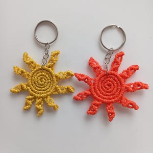May include: Two sun-shaped keychains crafted from knotted cord. One keychain is yellow and the other is orange. Each features a central spiral design with radiating, textured rays and is attached to a silver-toned metal split ring by a short chain.