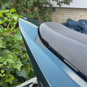 Może przedstawiać: Zbliżenie na turkusowe siedzenie motocykla i tylny błotnik. Siedzenie jest czarne i teksturowane, kontrastując z gładkim, metalicznym wykończeniem błotnika. Tło przedstawia zielone liście i ceglany mur.