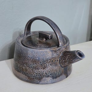 May include: A ceramic teapot with a textured, mottled finish in shades of dark brown, gray, and metallic bronze. The teapot features a wide, tapered body, a short spout, a flat lid with a small rectangular handle, and a high, arched handle.