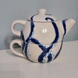 May include: A ceramic tea-for-one set consisting of a stackable teapot and teacup. The off-white glazed surface features a hand-painted pattern of bold, dark blue swirling lines that wrap around both the teapot and the cup, creating a cohesive design.