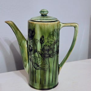 May include: A tall, cylindrical ceramic teapot with a glossy, variegated green glaze. The body features black line-art illustrations of lotus flowers and lily pads. It includes a matching lid with a small knob, a curved spout, and a handle.