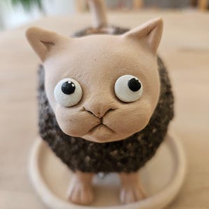 Op de afbeelding: Een keramieken kattenbeeldje met een gladde lichtbeige kop en een getextureerd donkerbruin lichaam. De kat heeft grote, ronde witte ogen met zwarte pupillen, puntige oren en een kleine staart op een bijpassende ronde beige schotel.