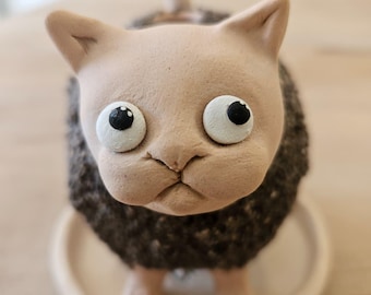 Puede incluir: Una figura de gato de cerámica con cabeza de color beige claro liso y cuerpo de color marrón oscuro texturizado. El gato tiene ojos grandes, redondos y blancos con pupilas negras, orejas puntiagudas y una cola pequeña sobre una base.
