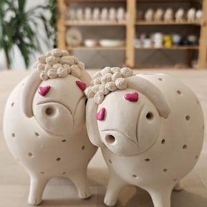 Peut inclure: Deux figurines de moutons en céramique. Les moutons sont blancs avec des dessus texturés en forme de laine et des accents roses en forme de cœur. Chaque mouton a de petits détails en points foncés et un design fantaisiste et amical. Les figurines sont sur une surface claire.