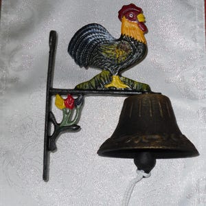 May include: A cast iron bell shaped like a rooster, with a black bell and a decorative bracket. The rooster is painted with yellow, red, and black details. The bracket features painted flowers. A white cord hangs from the bell.