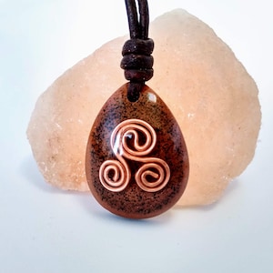 May include: A teardrop-shaped pendant with a copper spiral design, suspended from a dark brown leather cord. The pendant has a speckled brown and orange pattern. The background features a light orange, textured stone.