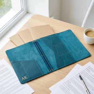 May include: A teal leather journal cover featuring an open interior with multiple pockets and elastic bands for holding notebooks. The bottom left pocket is embossed with the gold letters AK. The cover is laid flat, revealing its suede-like texture.