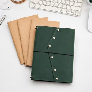 May include: A dark green notebook cover with a pointed flap secured by five silver metal studs and a black elastic band, accompanied by three light brown paper notebooks tucked behind it, all arranged on a clean white surface.