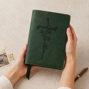 May include: A dark green journal with a smooth, matte cover featuring an embossed cross design intertwined with floral vines. The rectangular notebook includes a matching green ribbon bookmark extending from the bottom edge.