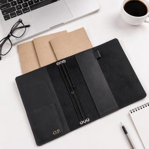May include: A black leather notebook cover featuring an elastic cord binding system, interior pockets, and a pen loop. The cover is personalized with the gold-embossed initials O.P. on the bottom left pocket, designed to hold multiple paper inserts.