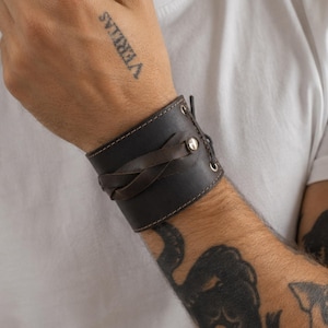 Könnte beinhalten: Ein dunkelbraunes Lederarmband mit weißen Nähten und einem geflochtenen Lederakzent. Das Armband hat Metallösen und einen Knopfverschluss. Das Wort "VERITAS" ist auf der Hand der Person tätowiert.