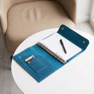Könnte beinhalten: Offener teal-farbener Lederordner mit silbernem Ringmechanismus, einem Stift und einem Stapel weißem Papier. Eine Kreditkarte befindet sich in einer Tasche auf der linken Seite. Der Ordner ist auf einem weißen Tisch geöffnet.