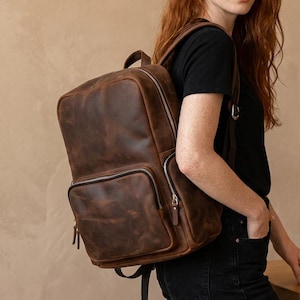 May include: A dark brown leather backpack featuring a large main compartment and a smaller front zippered pocket. The bag has adjustable shoulder straps and a structured rectangular shape with visible stitching and a smooth, matte finish.