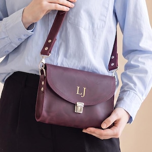 Puede incluir: Un bolso bandolera de cuero morado oscuro con cierre de solapa, un broche de metal plateado y una correa ajustable. La parte frontal del bolso presenta las letras del monograma LJ en color dorado centradas en la solapa, ofreciendo un diseño limpio.