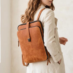 Könnte beinhalten: Ein brauner Lederrucksack mit rechteckiger Form und Klappenverschluss. Der Rucksack hat einen Reißverschluss an der Seite und verstellbare Träger. Das Leder hat ein strukturiertes Aussehen. Der Rucksack ist zum Tragen auf dem Rücken konzipiert.