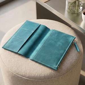 Puede incluir: Una cartera o funda para cuaderno de cuero color verde azulado con acabado mate liso, costuras blancas en contraste, múltiples ranuras para tarjetas en el interior izquierdo y un compartimento con cremallera a la derecha con tirador a juego.