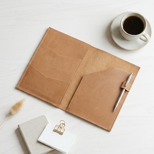 Puede incluir: Una funda de cuero color canela para cuaderno con bolsillos interiores y un bolígrafo plateado en un lazo lateral. Cerca hay un cuaderno gris pequeño, una tarjeta blanca con un clip de metal dorado, un tallo de planta seco y café negro.