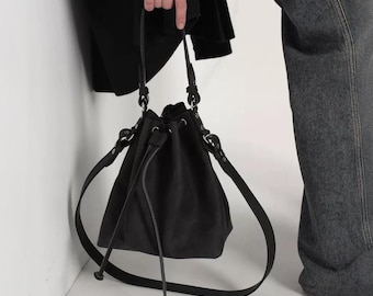 May include: A black bucket bag with a drawstring closure and long shoulder straps. The bag is made of a soft material and has a simple, minimalist design. The bag is held by a person wearing a black shirt and gray jeans.
