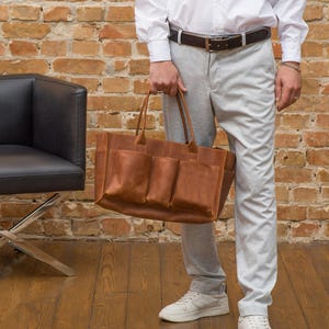 K&ouml;nnte beinhalten: Eine braune Ledertasche mit mehreren Taschen. Die Tasche hat einen oberen Griff und wird von einer Person gehalten.