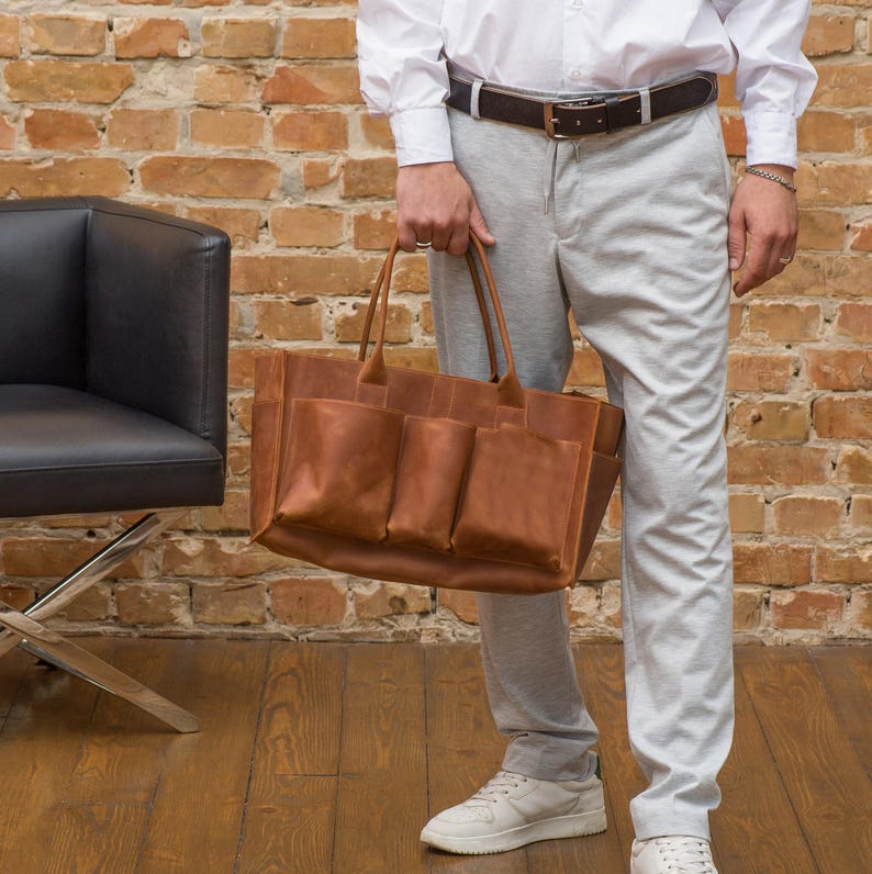 K&ouml;nnte beinhalten: Eine braune Ledertasche mit mehreren Taschen. Die Tasche hat einen oberen Griff und wird von einer Person gehalten.