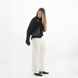 K&ouml;nnte beinhalten: Eine schwarze Umh&auml;ngetasche mit rechteckiger Form. Die Tasche wird &uuml;ber der Schulter getragen. Die Person auf dem Bild tr&auml;gt ein schwarzes Langarmshirt und eine wei&szlig;e Hose. Der Hintergrund ist wei&szlig;.