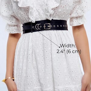 May include: A black leather belt, 2.4 inches (6 cm) wide, with a silver-colored buckle and eyelets. The belt is worn over a white dress with eyelet detailing. The belt is a waist cincher.