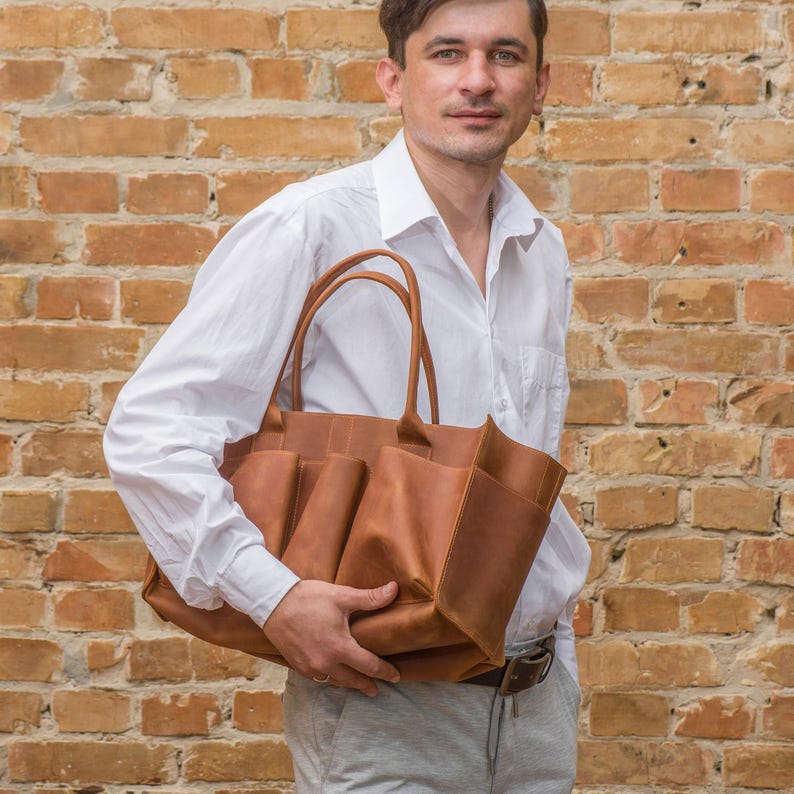 K&ouml;nnte beinhalten: Eine braune Ledertasche mit mehreren Taschen und einer gro&szlig;en &Ouml;ffnung. Die Tasche wird von einer Person getragen, die ein wei&szlig;es Hemd und eine graue Hose tr&auml;gt.