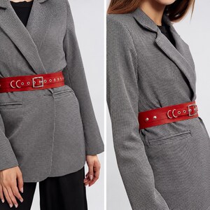 May include: A red leather belt with a silver buckle and rivets. The belt is worn over a gray and black houndstooth blazer. The belt is approximately 2 inches wide.
