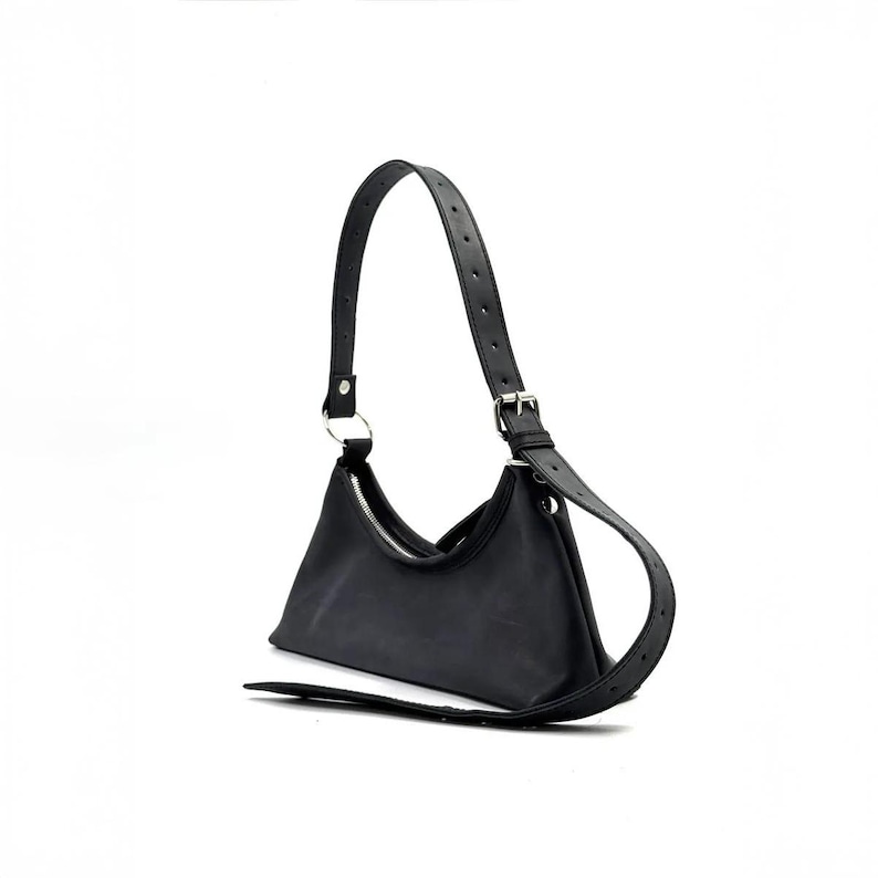 May include: Black shoulder bag with a crescent shape. The bag features a silver zipper, a long adjustable strap, and a silver metal ring. The bag is made of a smooth material.