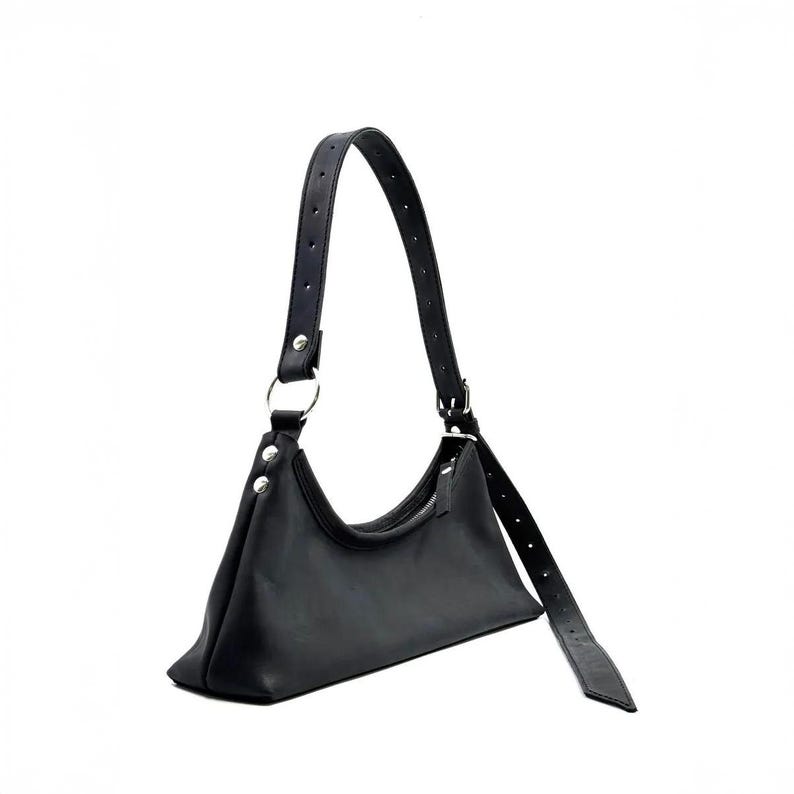 May include: Black shoulder bag with a curved top and a long, adjustable strap. The bag features a silver metal ring and rivets attaching the strap to the body. The bag is made of a smooth, matte material.
