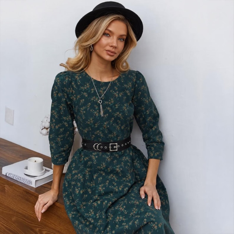 May include: A dark green floral print dress with long sleeves and a black belt. The dress has a round neckline and is accessorized with a silver necklace. A black hat is worn on the head.