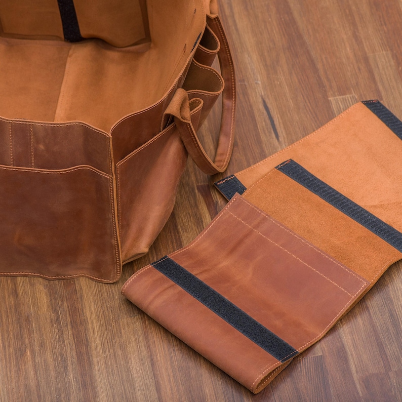 K&ouml;nnte beinhalten: Eine braune Ledertasche mit mehreren Taschen und einem schwarzen Riemen. Die Tasche ist offen und das Futter ist sichtbar. Die Tasche liegt auf einer Holzoberfl&auml;che. Neben der Tasche liegt ein separates St&uuml;ck braunes Leder mit einem schwarzen Riemen.