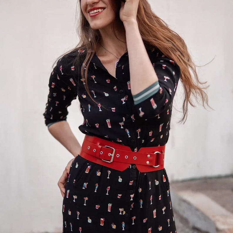 May include: A black button-down dress with a cocktail print, paired with a wide red belt. The belt has a silver buckle and multiple eyelets. The dress has long sleeves with a green and white striped trim.