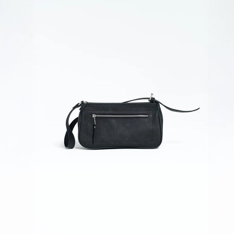 K&ouml;nnte beinhalten: Eine schwarze, rechteckige Umh&auml;ngetasche mit Rei&szlig;verschluss und einem langen, d&uuml;nnen Riemen. Die Tasche hat eine kleine, rechteckige Tasche auf der Vorderseite und einen silbernen Rei&szlig;verschluss. Die Tasche ist aus mattem Material.