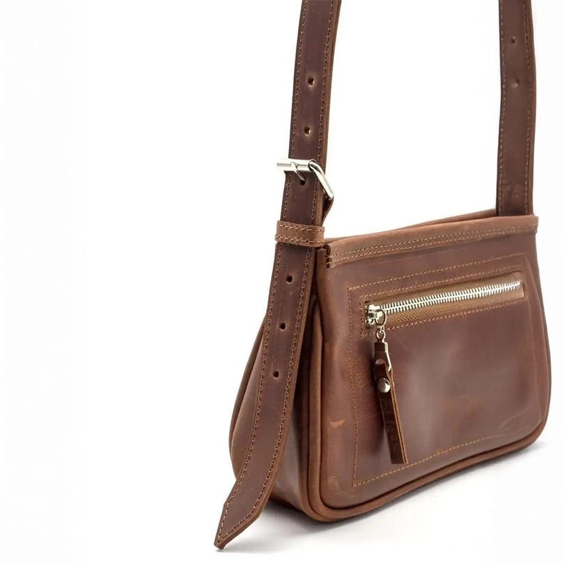 K&ouml;nnte beinhalten: Braune Leder-Umh&auml;ngetasche mit silbernem Rei&szlig;verschluss und verstellbarem Riemen. Die Tasche hat eine rechteckige Form mit einer Vordertasche und einem Leder-Rei&szlig;verschluss. Der Riemen ist mit einer silbernen Schnalle befestigt.