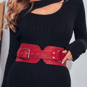 May include: A wide, red leather belt with a silver buckle and two silver rings. The belt has several small silver rivets and is worn over a black long-sleeved dress. The belt is a bold fashion accessory.