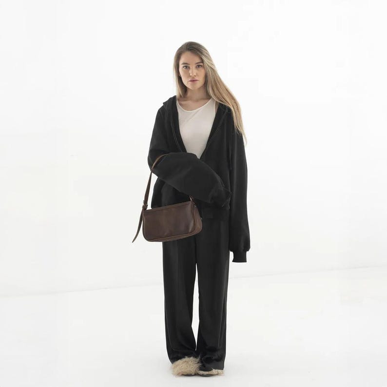 K&ouml;nnte beinhalten: Eine Frau tr&auml;gt einen schwarzen Kapuzenpullover, eine schwarze Hose und ein wei&szlig;es Oberteil. Sie h&auml;lt eine braune Leder-Umh&auml;ngetasche. Das Outfit ist l&auml;ssig und bequem. Die Tasche hat einen langen Riemen.