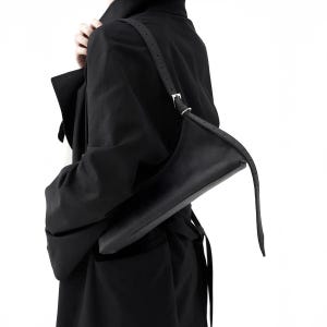 May include: A black shoulder bag with a long, adjustable strap. The bag has a sleek, minimalist design with a rectangular shape and a silver buckle. The bag is made of a smooth, matte material.