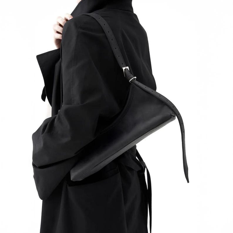 May include: A black shoulder bag with a long, adjustable strap. The bag has a sleek, minimalist design with a rectangular shape and a silver buckle. The bag is made of a smooth, matte material.