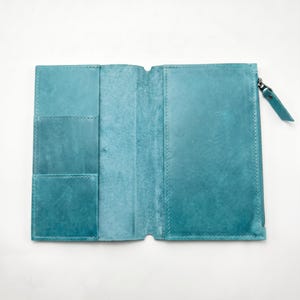 Op de afbeelding: Een opengevouwen blauwgroene lederen portemonnee met een gladde afwerking en zichtbare stiksels. De linkerkant bevat twee gestapelde kaartsleuven, terwijl de rechterkant is voorzien van een vak met ritssluiting en een bijpassende lederen trekker.