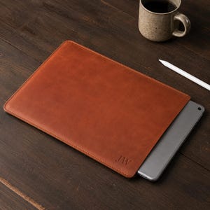 Op de afbeelding: Een bruine leren tablethoes met de initialen "JW" in de rechter benedenhoek. Een tablet is gedeeltelijk zichtbaar in de hoes. Een witte stylus en een koffiemok liggen op een donker houten oppervlak.