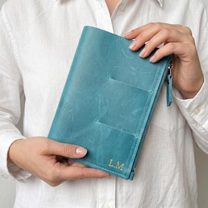 May include: A teal leather notebook cover featuring a vertical zippered pocket on the right side and the gold embossed initials L.M. at the bottom right corner. The cover has a smooth, matte texture with visible natural grain and stitching.