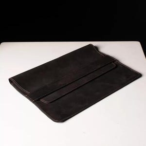 Könnte beinhalten: Eine dunkelbraune Laptop-Hülle aus Leder mit Klappenverschluss. Die rechteckige Hülle hat abgerundete Ecken und weiße Nähte. Das Leder hat eine strukturierte Optik, und die Klappe wird mit einem passenden Lederriemen befestigt.